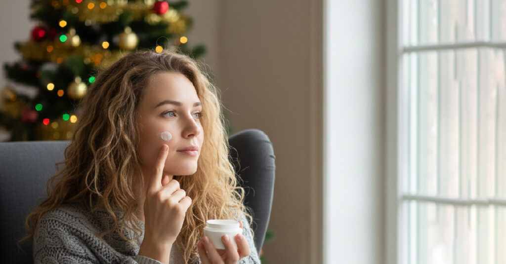 🎄My Christmas Glow: The Winter Skincare Routine That Saves My Skin Every Year "Woman applying moisturizer on her face during winter skincare routine near a window and Christmas tree."